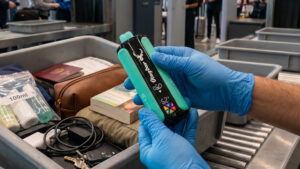 An airport security officer wearing blue gloves holds and inspects a Stagbar vape device over a plastic tray filled with travel items at a security checkpoint.